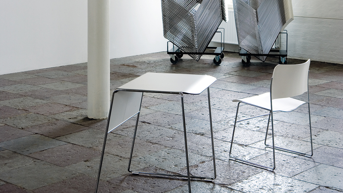 AnInstallation image of a white Tutor table and 40/4 chair in front, and stacks of Tutor tables and 40/4 chairs on dollies in back