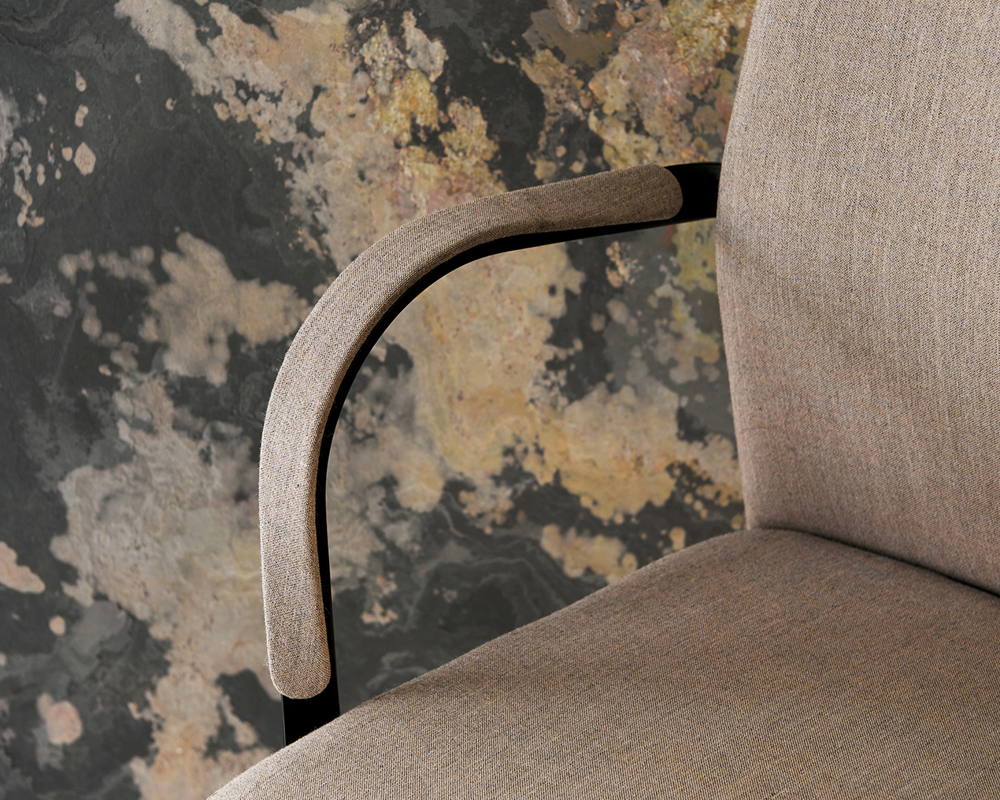 A closeup picture of an armrest of the MN1 chair with a beige upholstered seat against a patterned wall