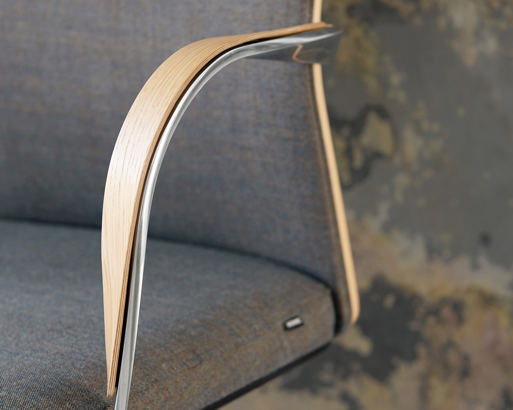 A closeup picture of an armrest of the MN1 chair with a beige upholstered seat against a patterned wall